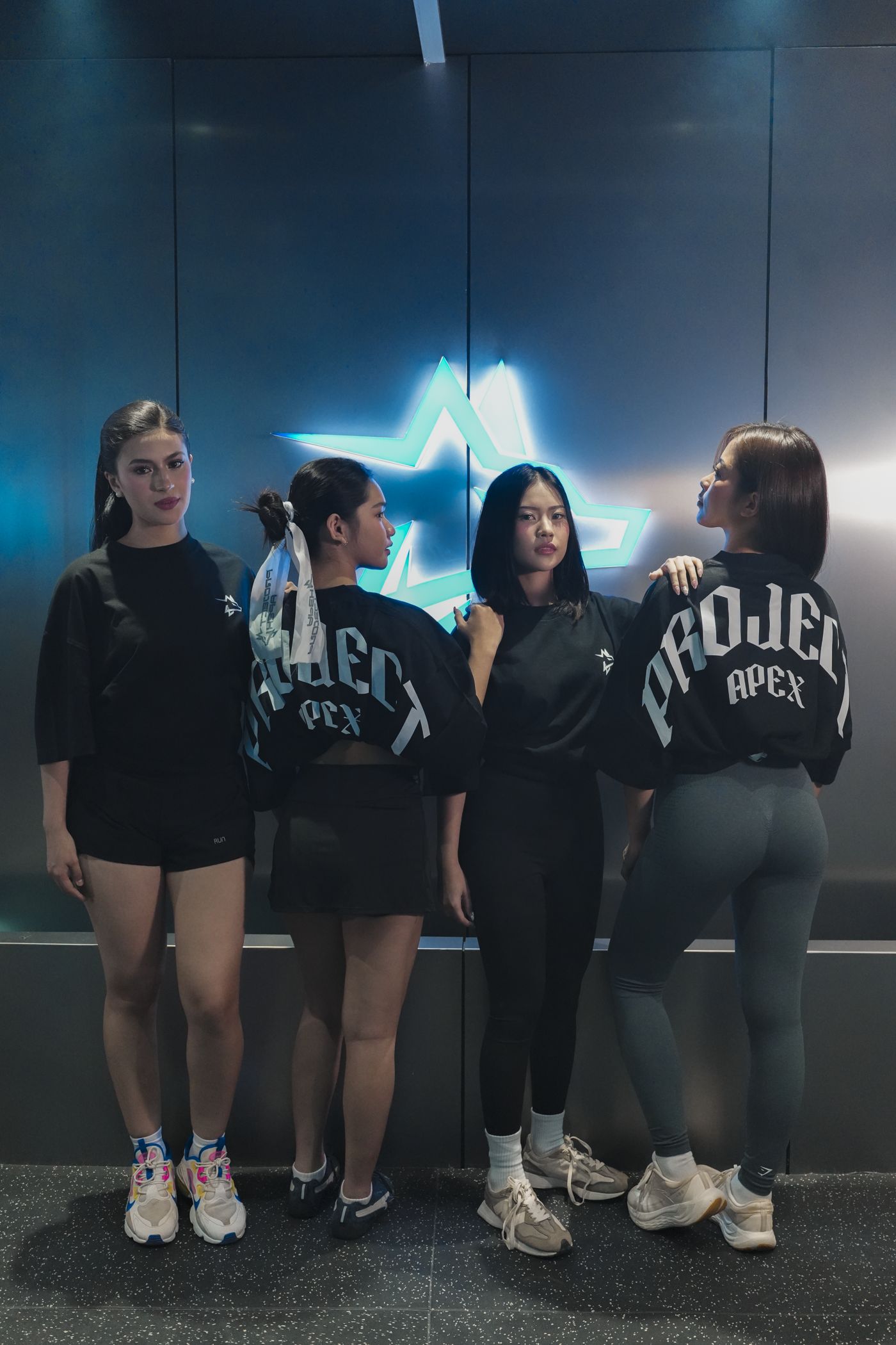 Four women in APX tees in front of the Project Apex neon wolf, two showing the arched back print